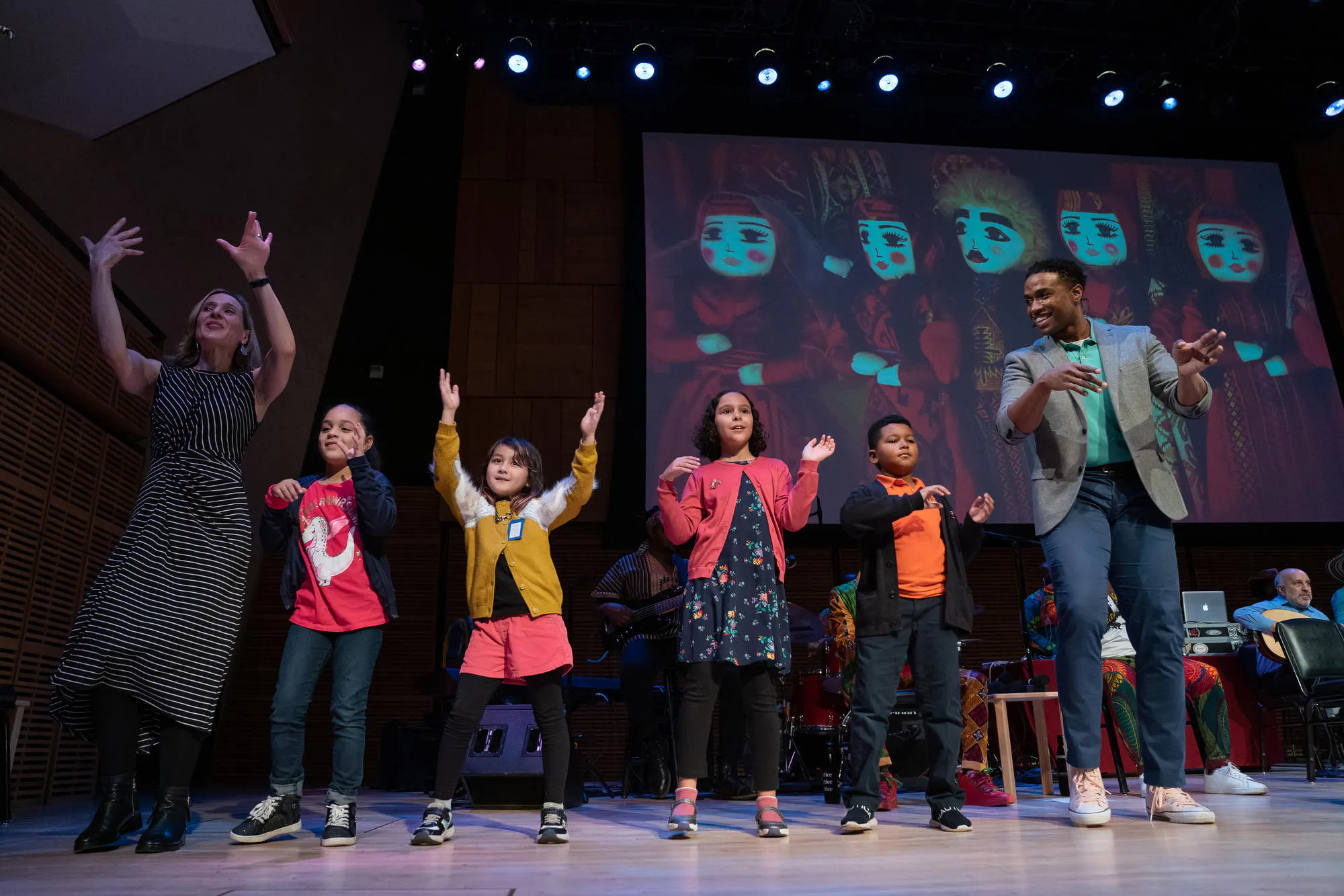 Four children dance on stage, guided by a man and a woman
