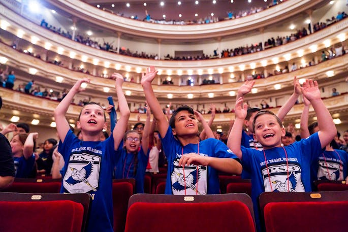 Children stand behind red velvet seats with their hands raised above their heads in Stern/Perelman.