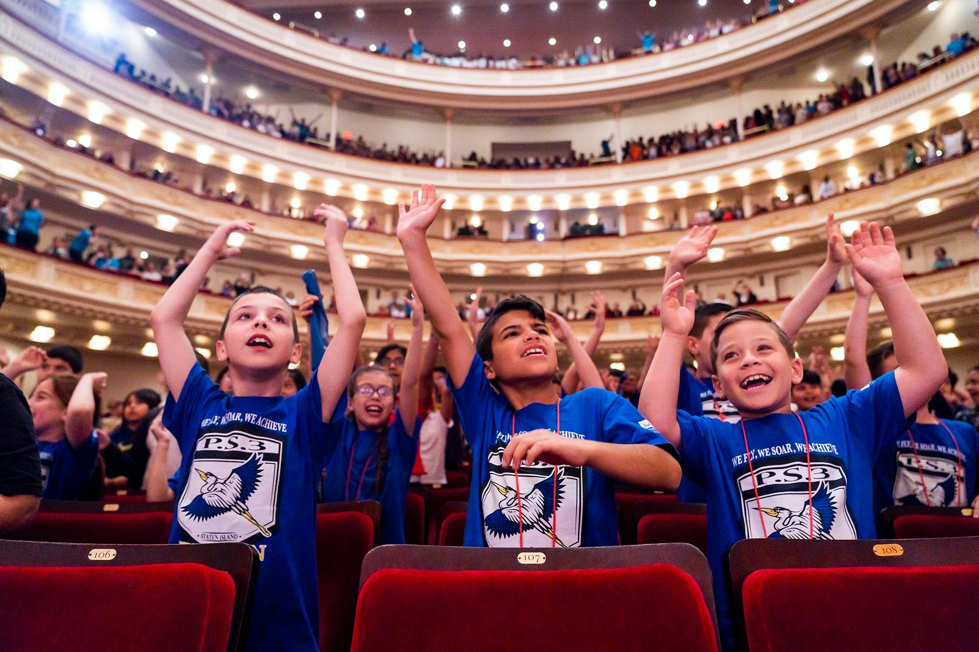 Children stand behind red velvet seats with their hands raised above their heads in Stern/Perelman.