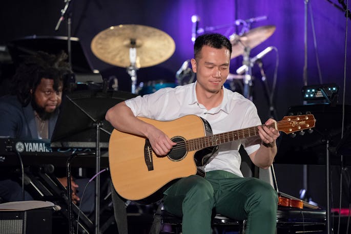 A man wearing a white polo and green pants smiles as he plays the guitar in front of a drum set.