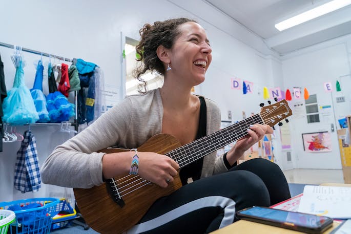 A young woman smiles playing the ukulele in a classroom.