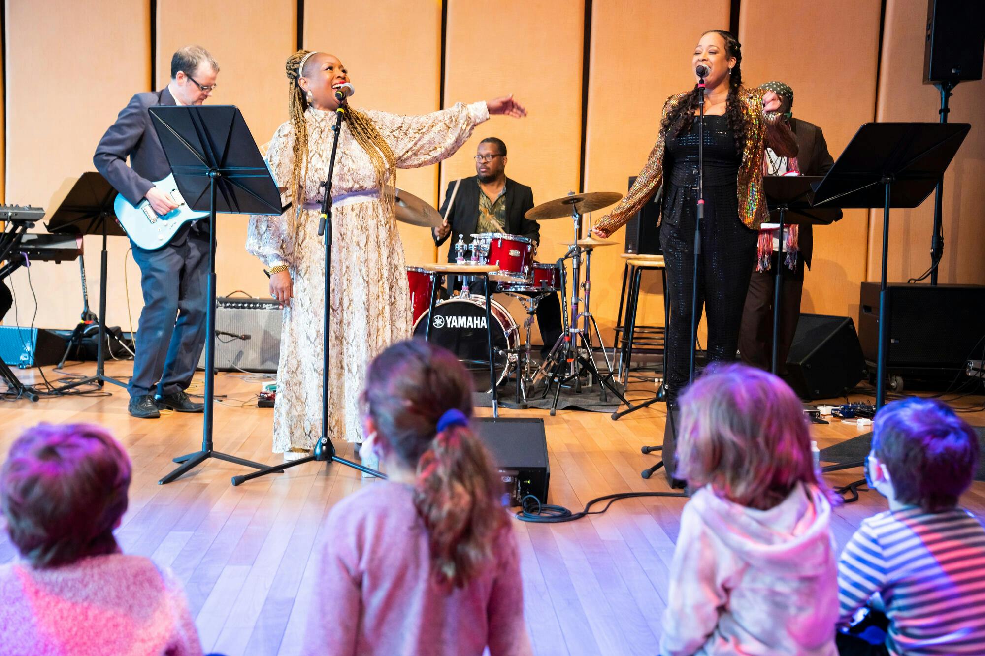 Children watch as two women sing, backed by a guitarist and drummer