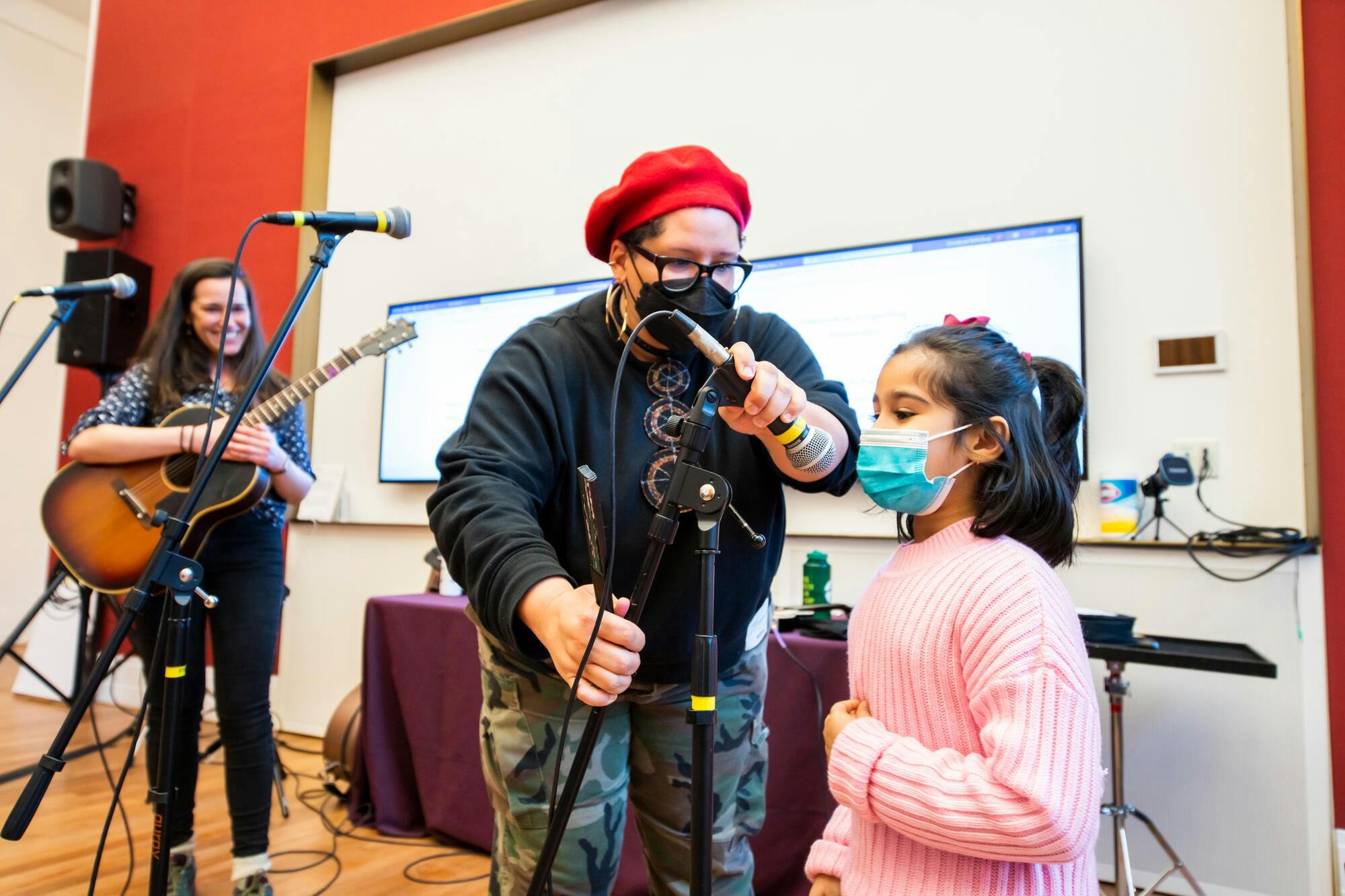 A woman in a red beret adjusts a microphone for a girl in a pink sweater