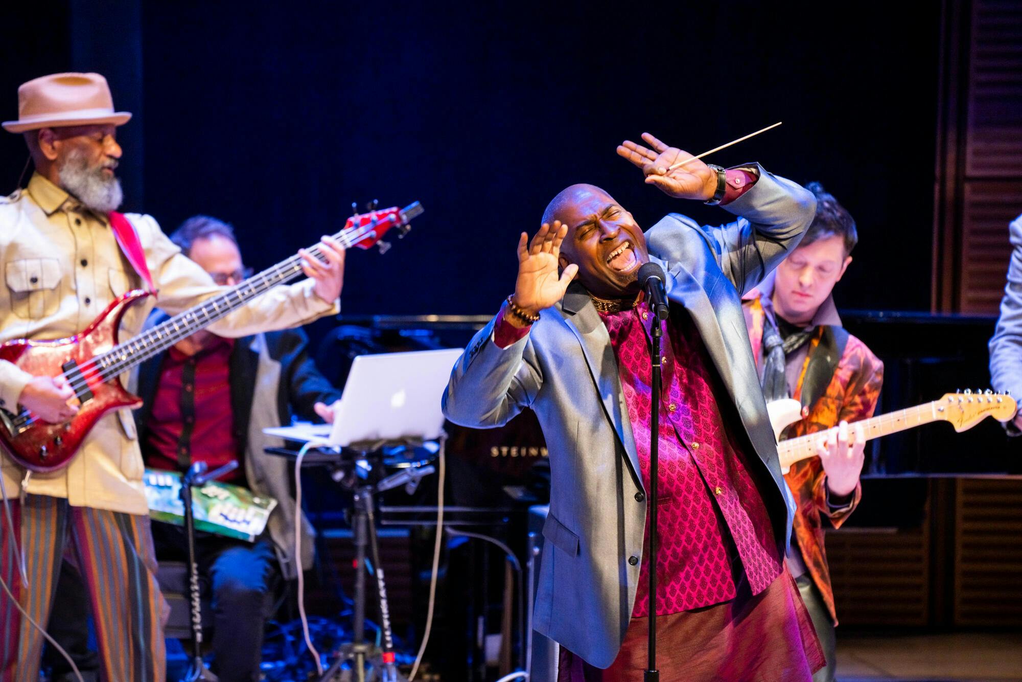 A man wails into a microphone, hands upraised, as a bassist plays