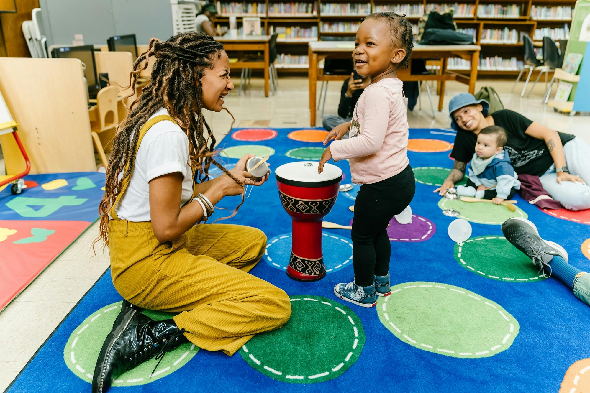 A woman smiles at a toddler standing over a drum on a colorful rug