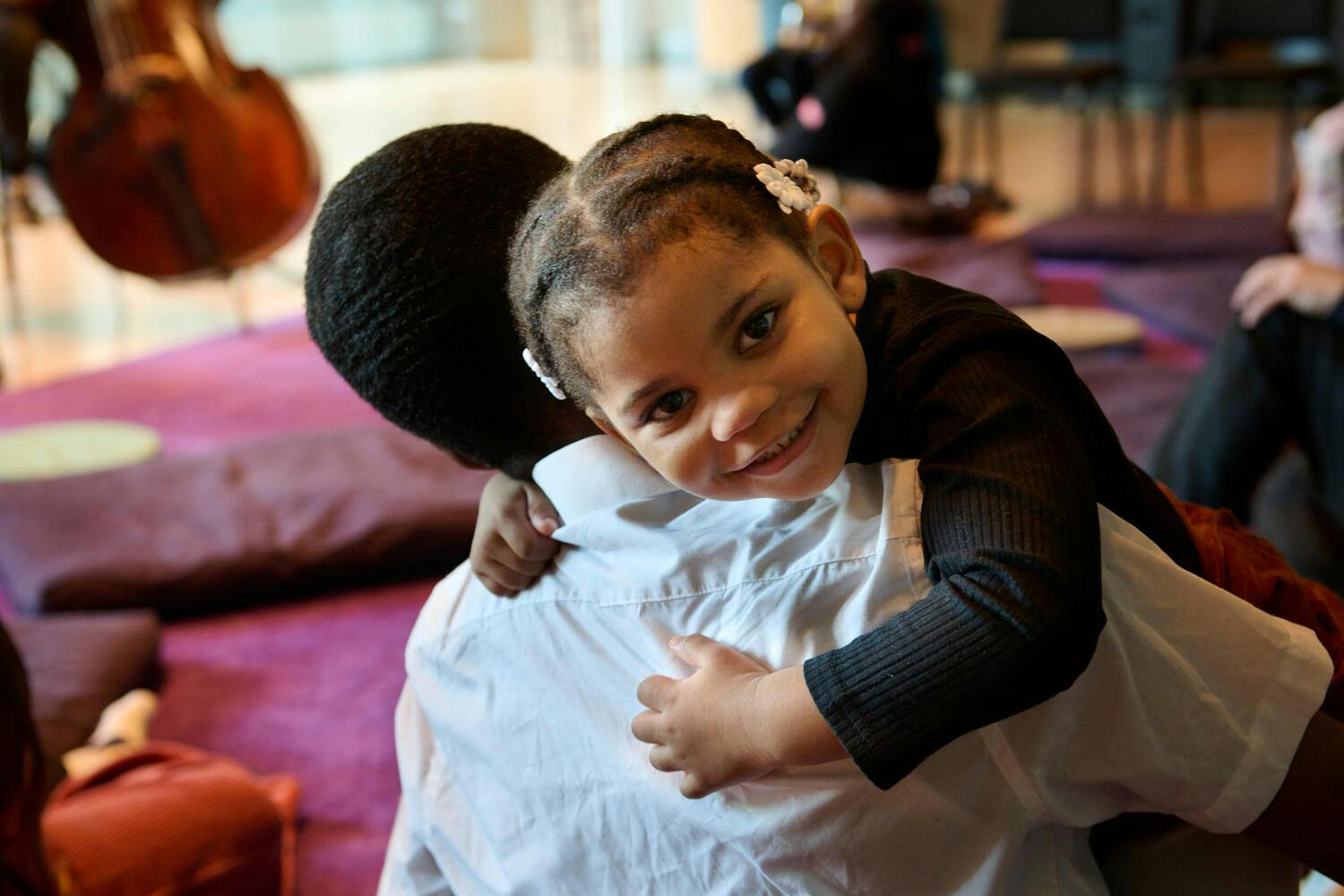 A toddler girl smiles at the camera, arms wrapped around her parent's neck