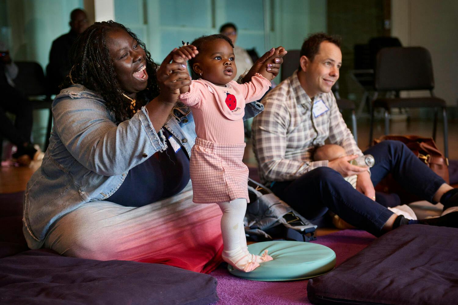 A smiling woman holds up her toddler girl as a man feeds a bottle to his baby