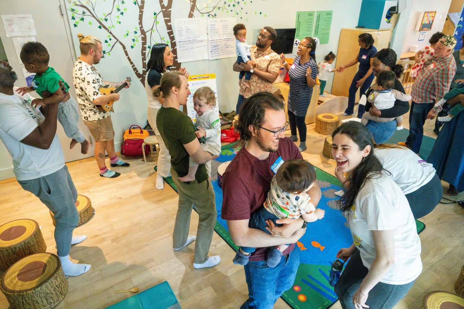 Parents hold their babies in a classroom as a man plays ukulele
