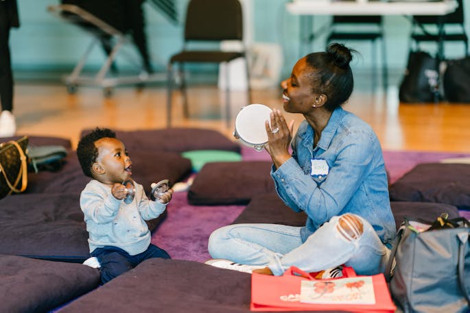 A smiling mother plays tambourine as her toddler watches