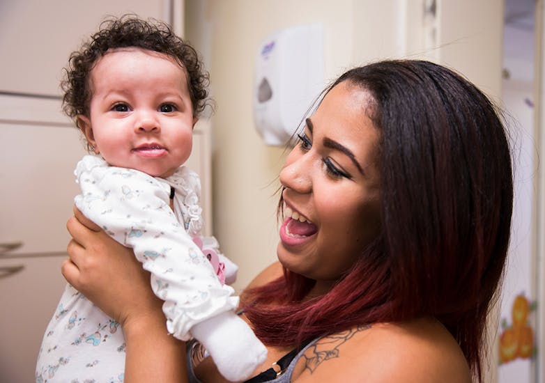 A woman with brown hair smiles as she holds up a baby with curly dark hair who looks at the camera.