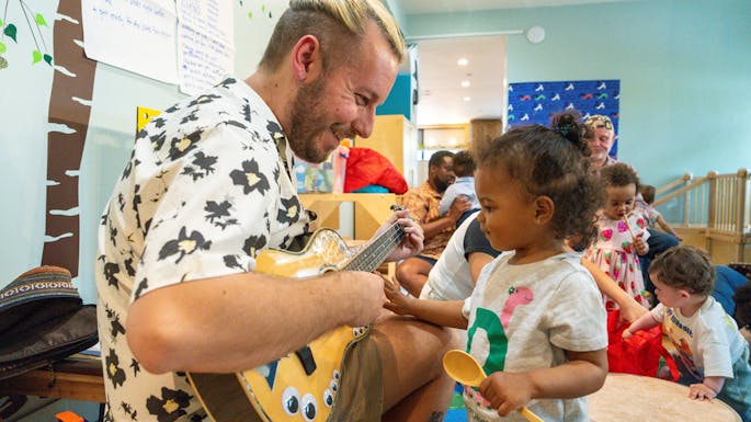 A man playing guitar smiles down at a toddler girl as she watches