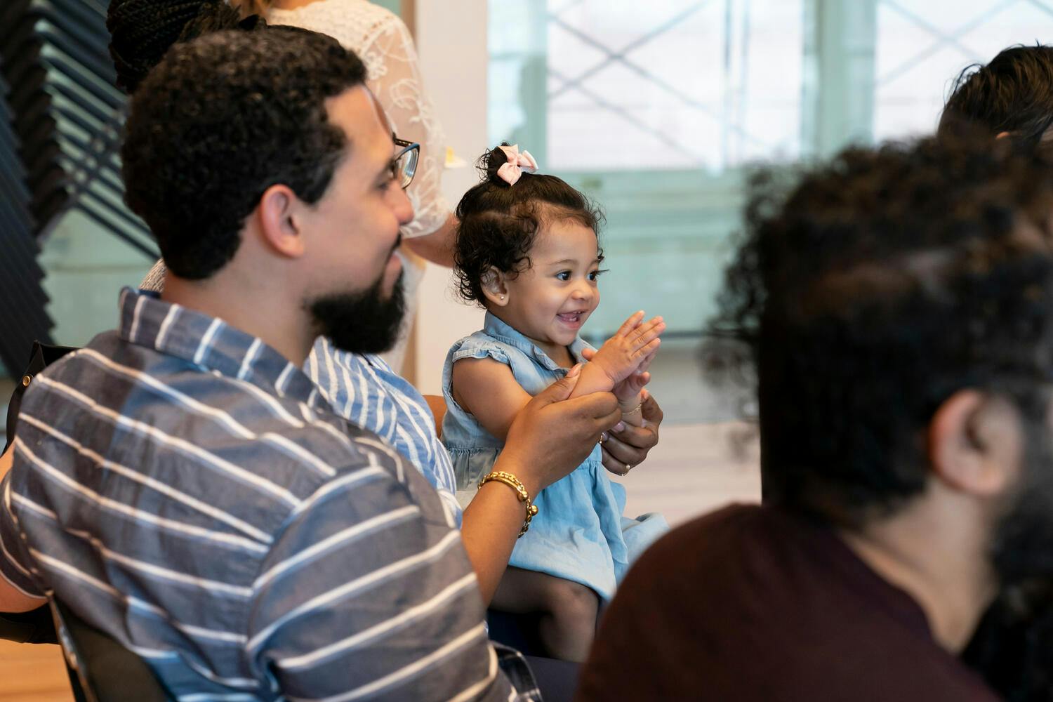 A smiling toddler claps, sitting in the lap of her parent
