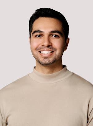 A young man with short, dark hair, dark eyes, light stubble, and a tan-colored light sweater smiles.
