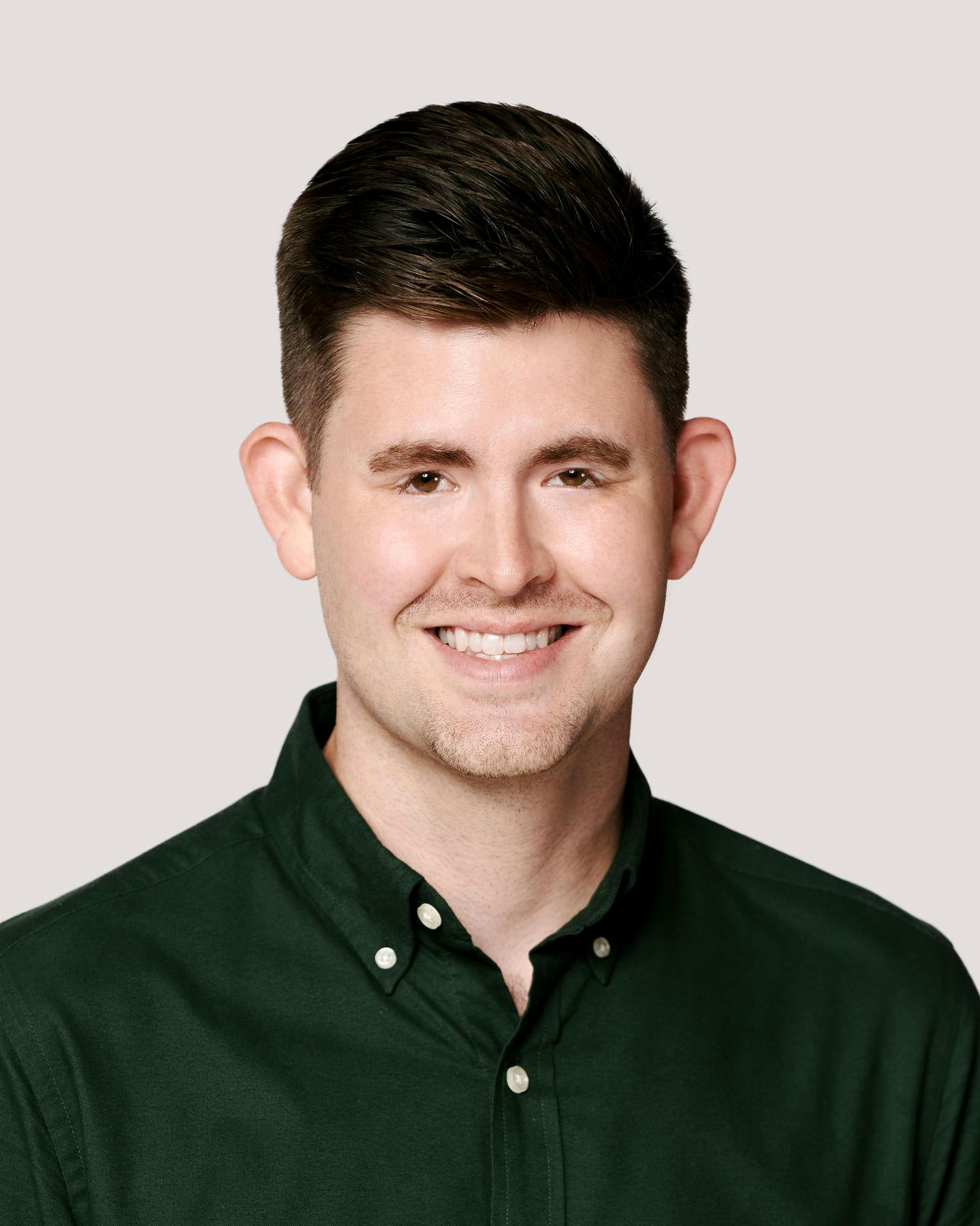 A young man in a green shirt with short, dark-blond hair and light facial hair smiles.