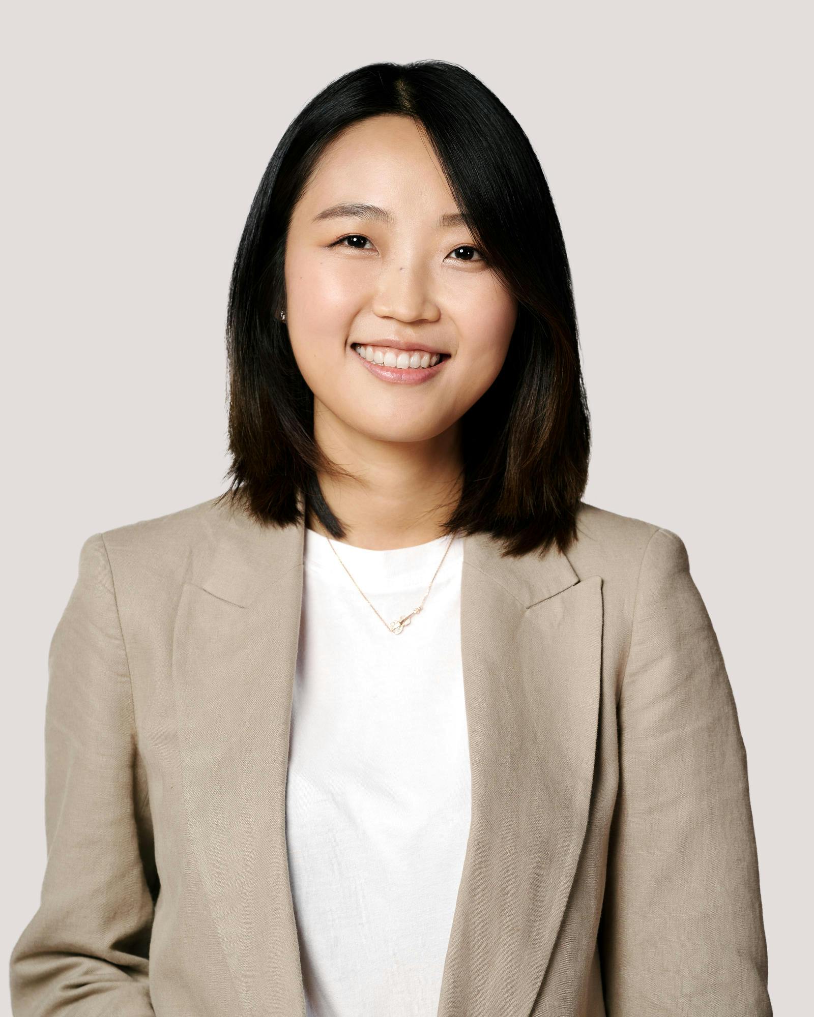 A young woman with dark hair and eyes wearing a light grey blazer and white Tshirt smiles.