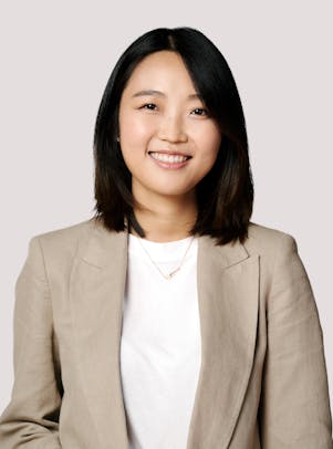 A young woman with dark hair and eyes wearing a light grey blazer and white Tshirt smiles.