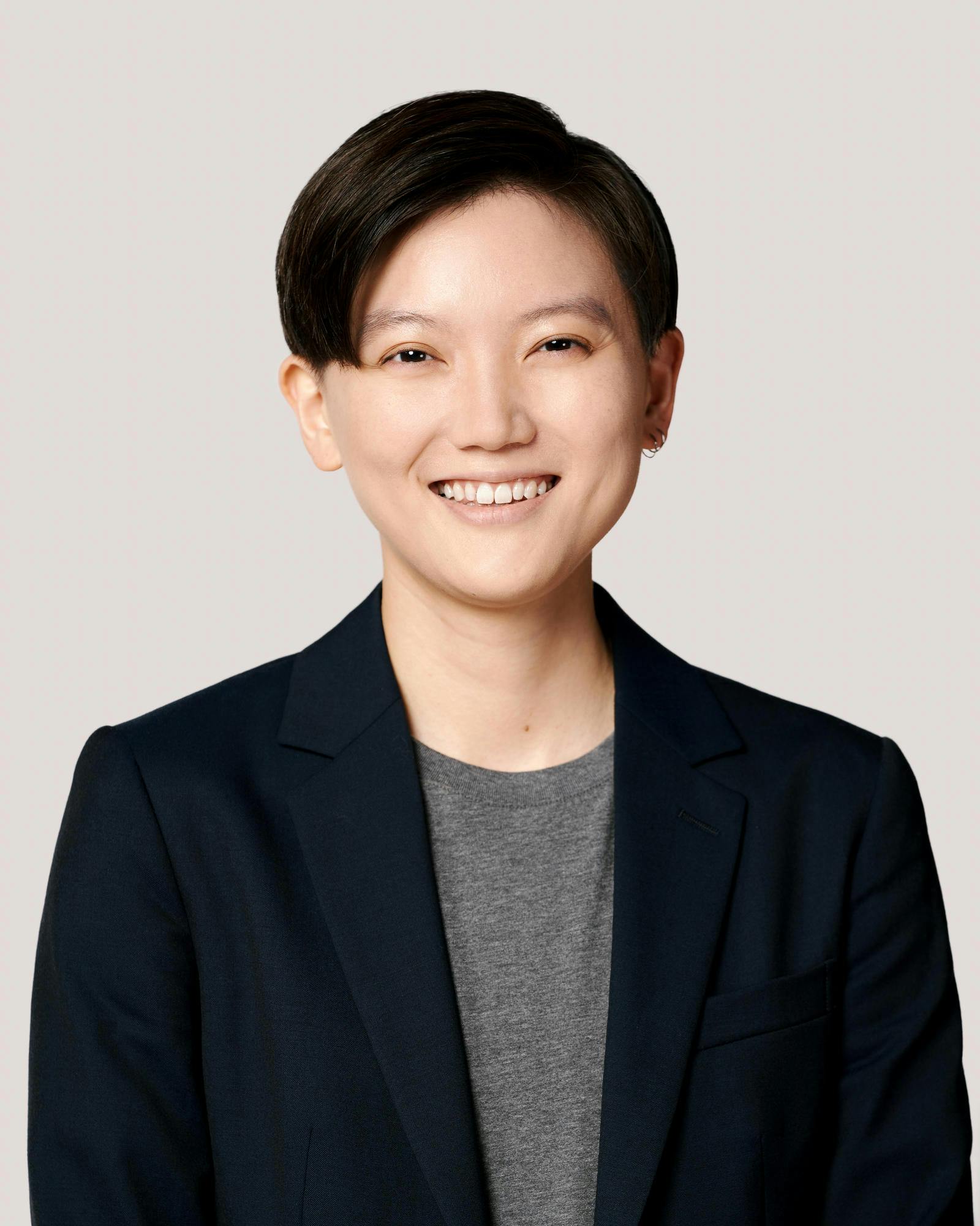 A young woman with short dark hair wearing a dark blazer and grey Tshirt, smiles.