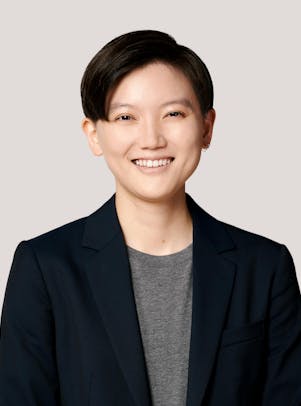 A young woman with short dark hair wearing a dark blazer and grey Tshirt, smiles.