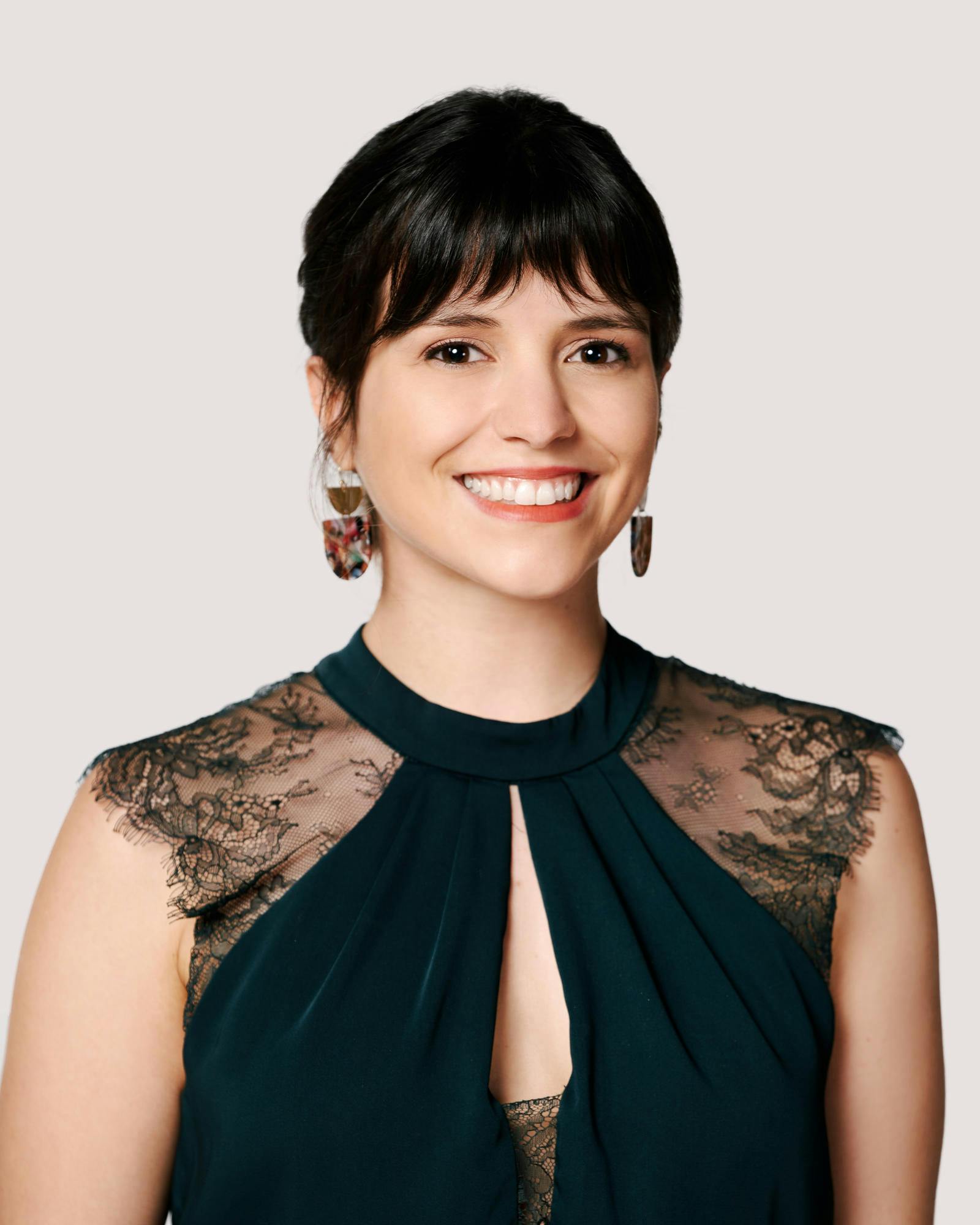 A young woman with dark hair and bangs, dark eyes, and a dark lace-adorned blouse smiles.