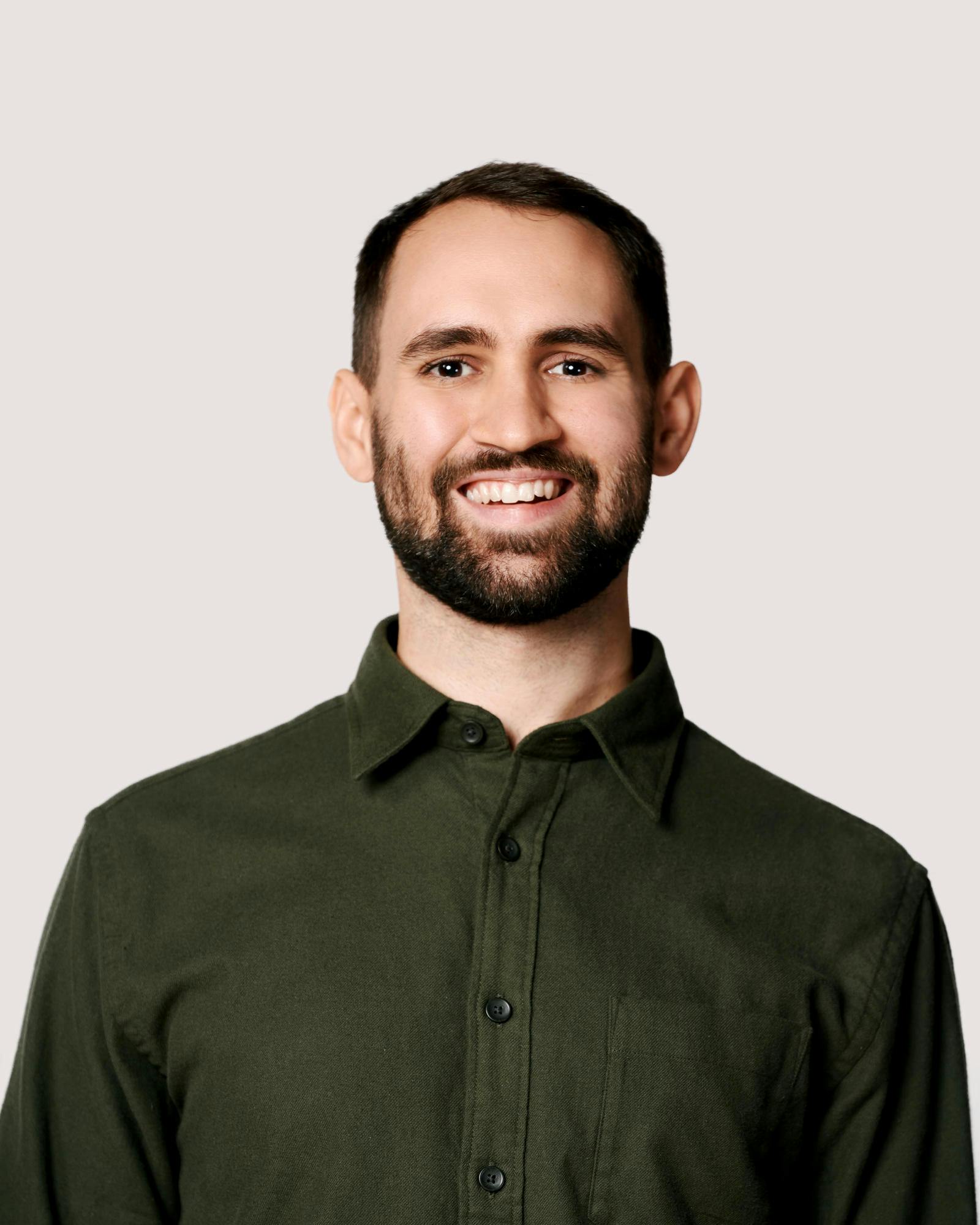 A young man in a green shirt with short, dark hair, dark eyes, and a short beard smiles.