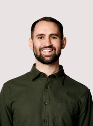 A young man in a green shirt with short, dark hair, dark eyes, and a short beard smiles.