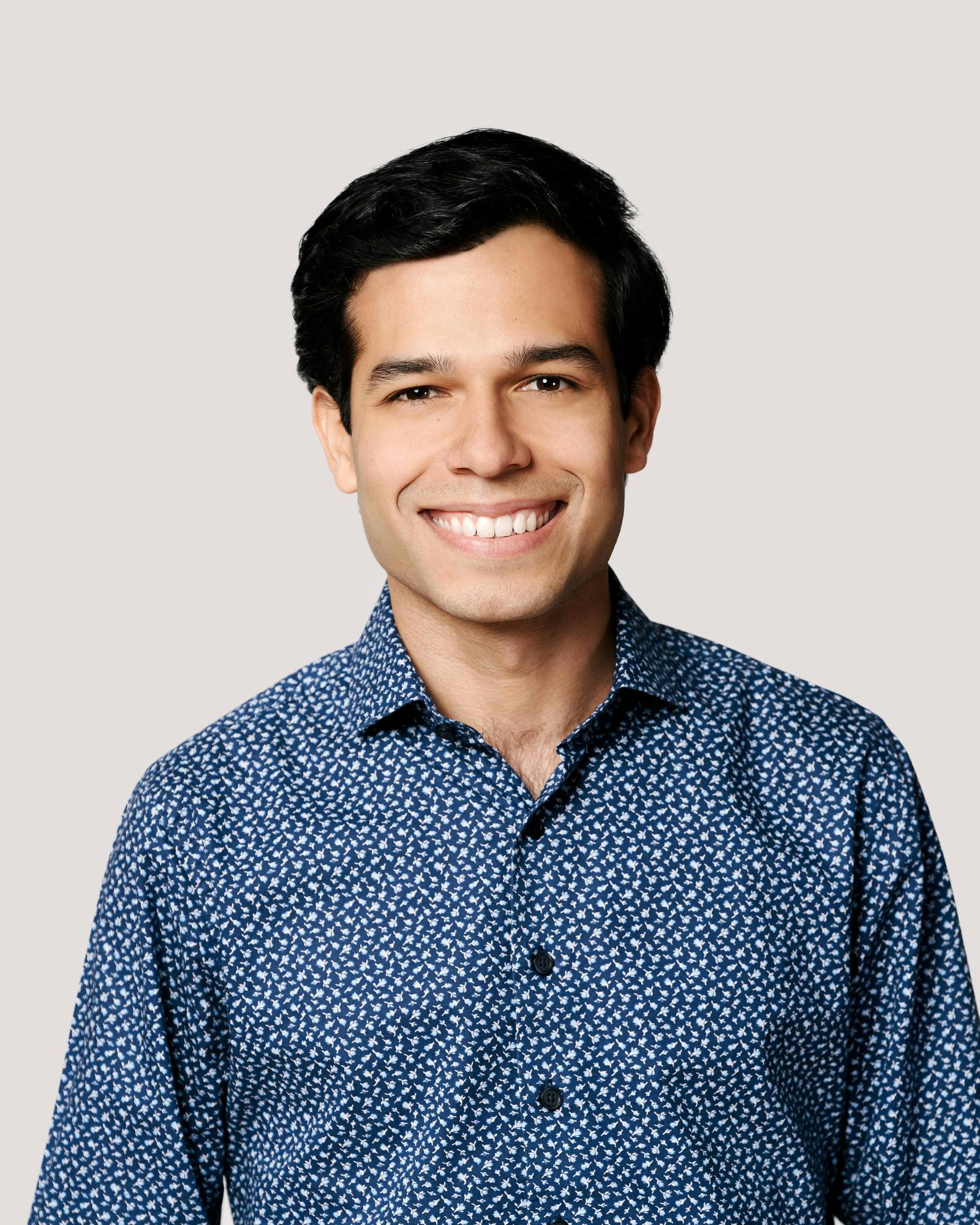 A young man in a blue printed button-up shirt with short dark hair and dark eyes smiles.
