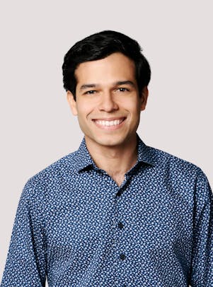A young man in a blue printed button-up shirt with short dark hair and dark eyes smiles.