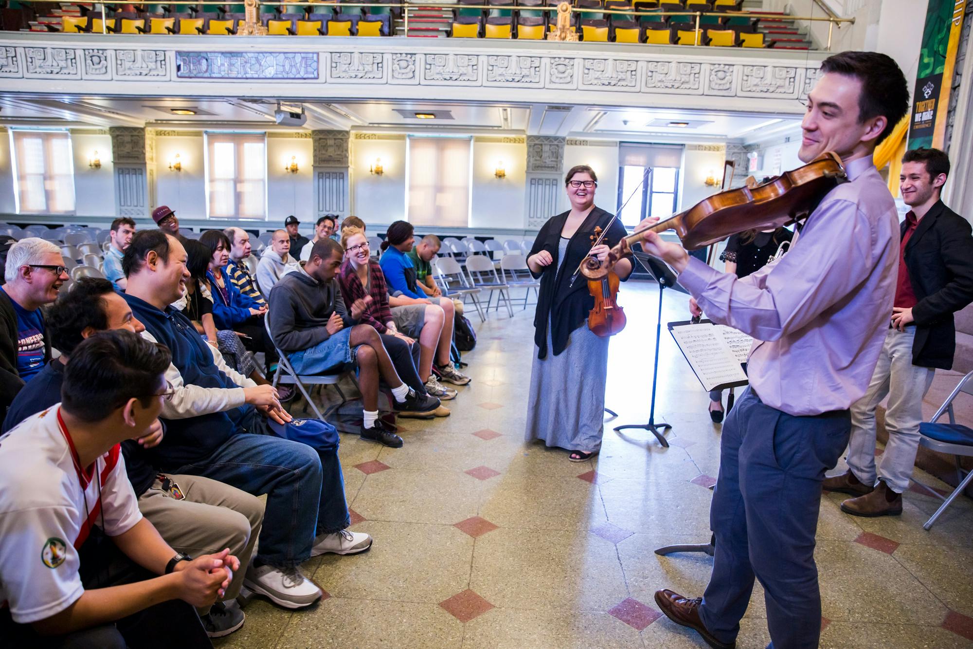 A man in formal attire smiles as he plays violin for an intimate seated audience.