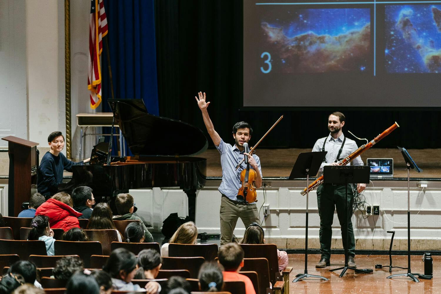 Students in a school auditorium watch as a young man with a violin holds up his hand