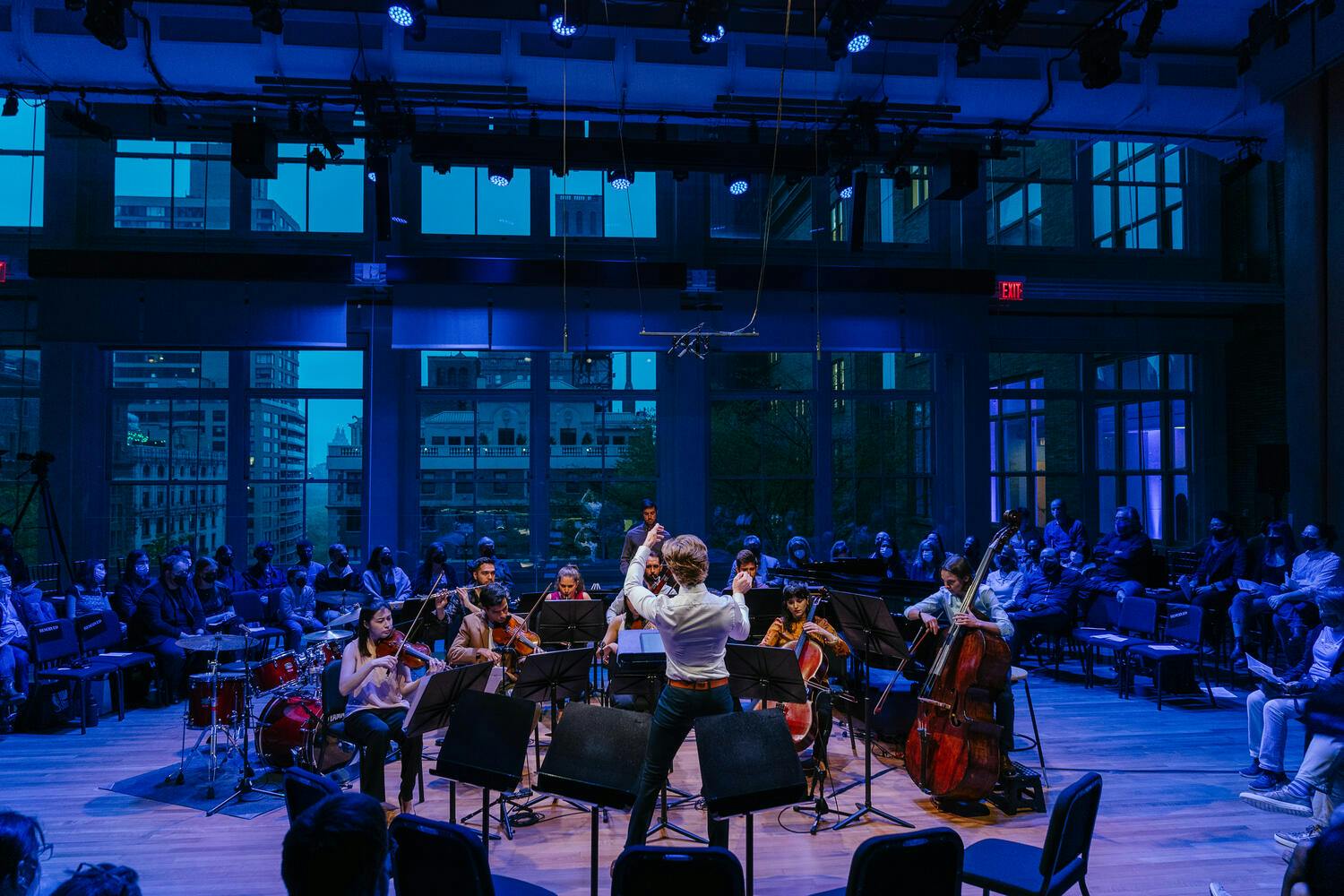 Surrounded by blue lighting, a conductor leads an ensemble encircled by an audience.