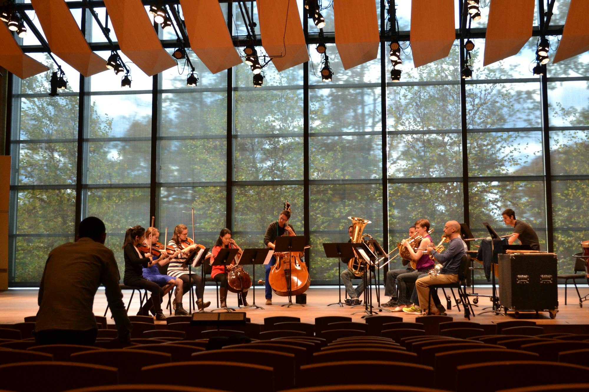 Ensemble Connect musicians rehearse on stage in front of a glass wall with trees in the background.