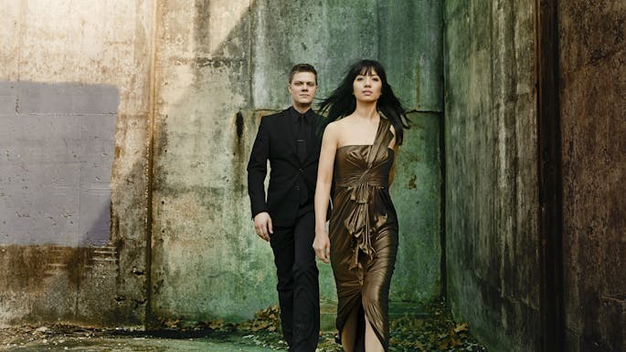 A young woman with dark hair wearing a brown formal gown walks in front of a man in a black suit.