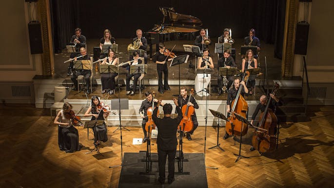 A group of musicians perform under a spotlight as a conductor leads them.