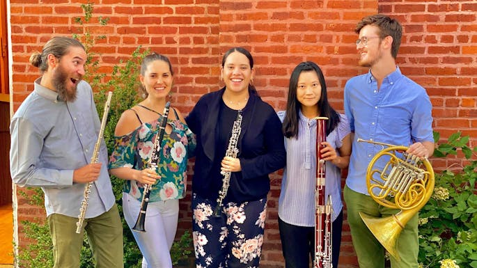 Five young musicians in casual attire hold instruments and stand against a brick wall.