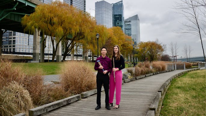 A young man holding timpani sticks poses on a wood walkway with a young woman holding a flute