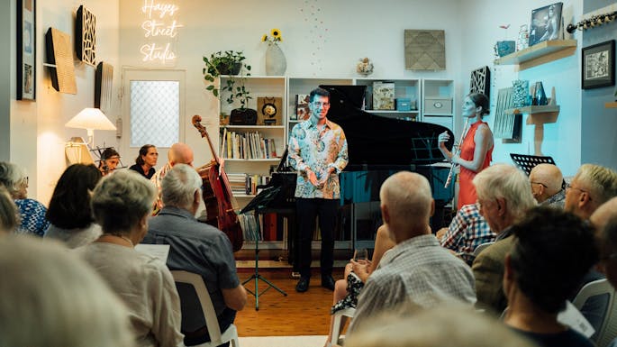 A young man speaks in a small room with a grand piano before a small audience