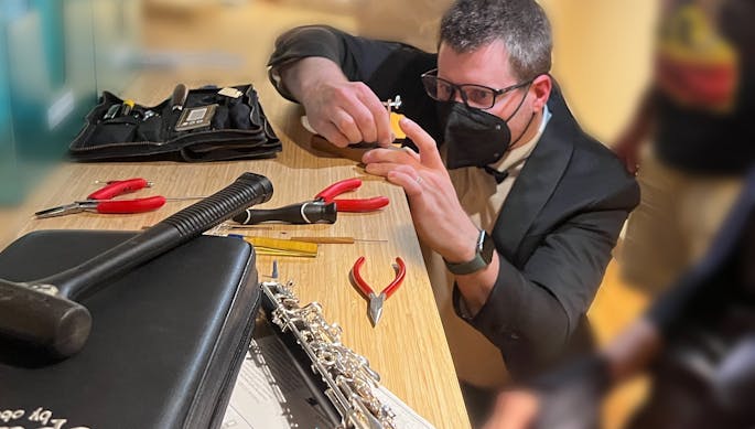A man in a blazer, glasses, and mask uses tools to work on an instrument.