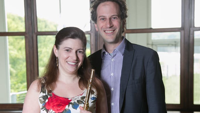 A woman and a man stand together behind a piano, in front of large windows. The woman holds a flute.