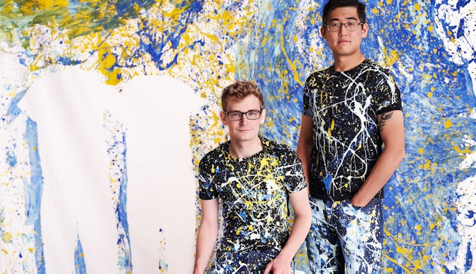 Two men, one sitting and one standing, are covered in yellow, white, and blue paint.