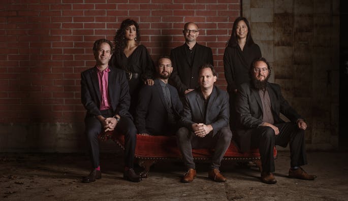 Seven people, four sitting and three standing, in black clothing pose together in front of a brick wall.