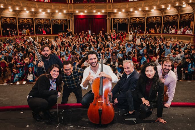 Seven musicians pose with instruments in front of a packed auditorium of children.