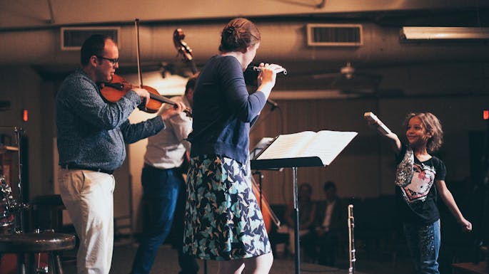 Three musicians perform together; in the foreground, a young girl smiles at them.