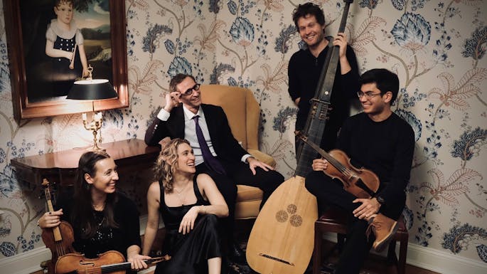 Smiling musicians of Theotokos pose with their instruments in a room with floral wallpaper