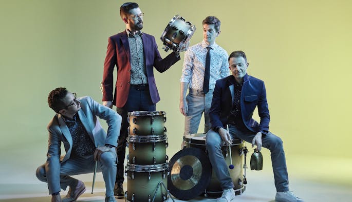 Four percussionists in blazers and patterned shirts against a green backdrop