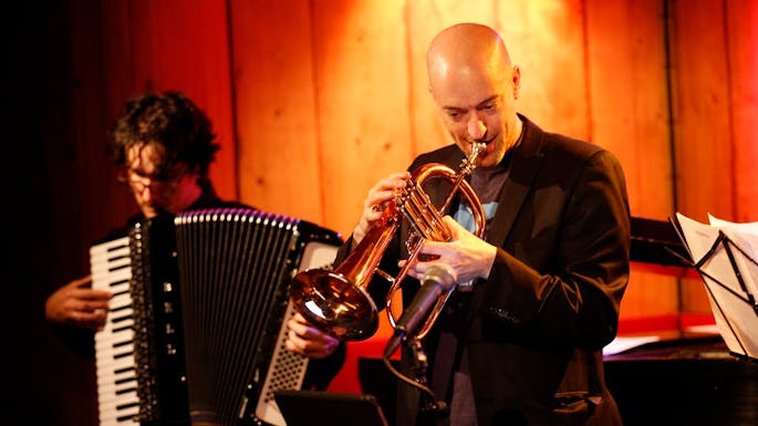 A bald man plays trumpet on stage as another man plays accordion behind him