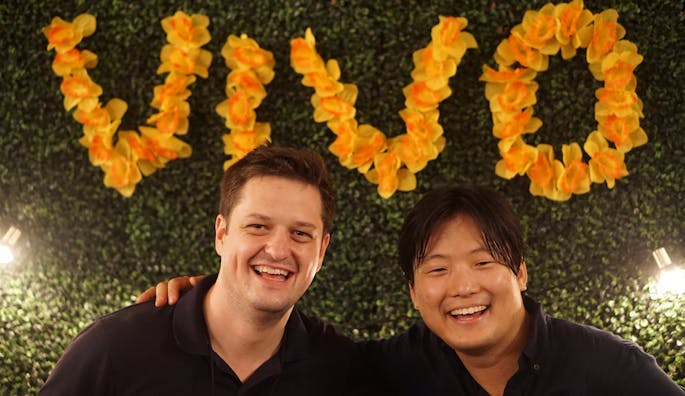 Two men stand smiling wearing black polos. “Vivo” is spelled in yellow flowers behind them.