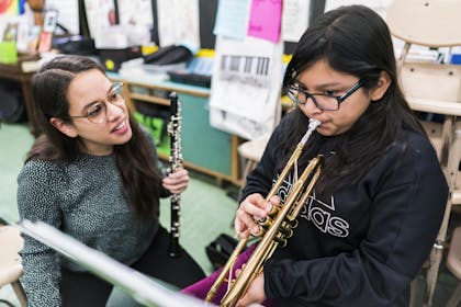 A female Ensemble Connect fellow works with a young girl playing the trumpet in a classroom.