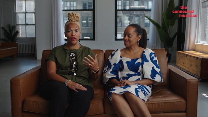 Monica Ellis and Toyin Spellman-Diaz sit on a couch speaking to camera.