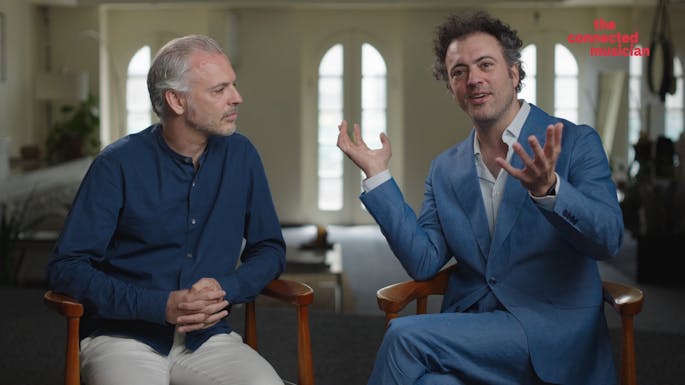 Eric Jacobsen and Colin Jacobsen sit together in tall chairs, Colin gesturing and smiling as he speaks to camera.