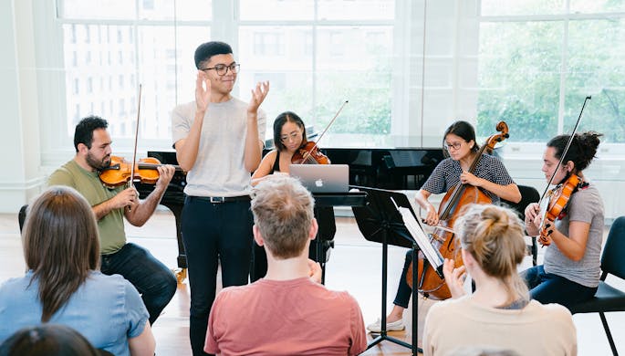 A string quartet plays as a young man in glasses claps in front of an audience.